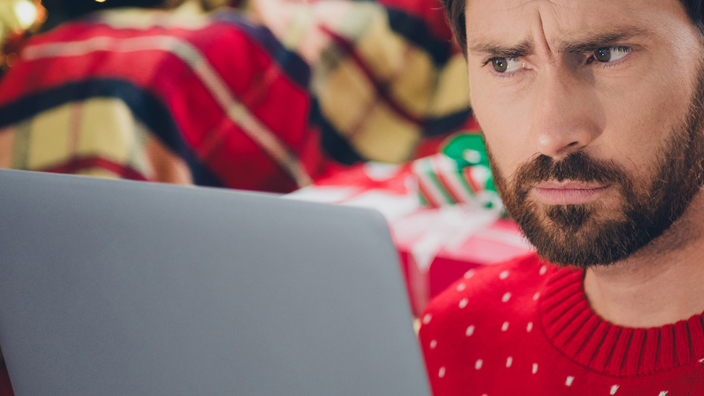 A man in an ugly holiday sweater looks at a laptop screen.