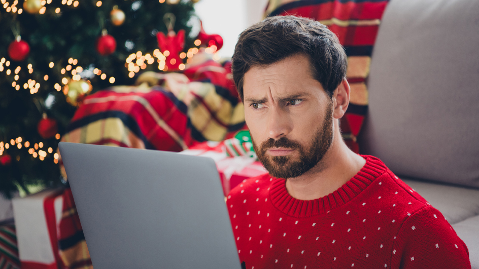 A man in an ugly holiday sweater looks at a laptop screen.