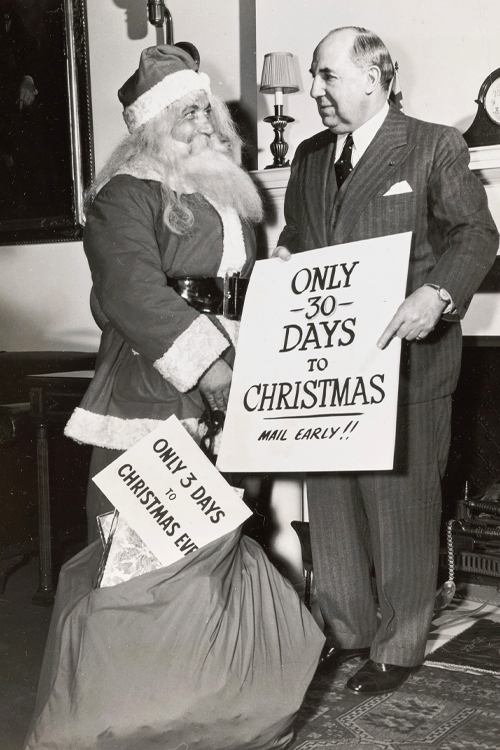 A black-and-white vintage photo of a man wearing a Santa Claus suit standing next to a man in a business suit