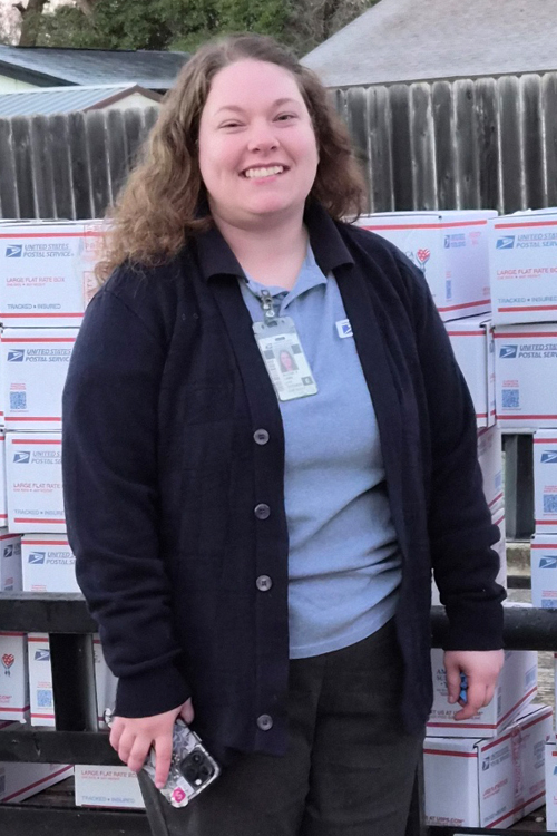 A woman in a postal uniform stands next to stacks of USPS-branded boxes