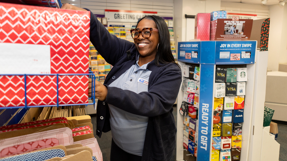 A smiling woman wearing a USPS retail clerk’s uniform adjusts a greeting card display in a Post Office retail lobby