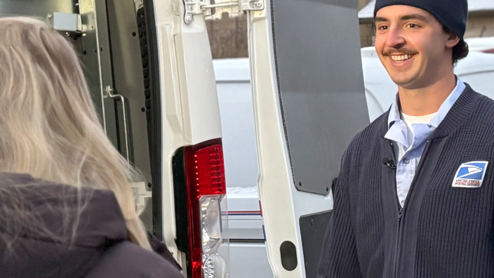 A smiling man wearing a Postal Service uniform stands next to a USPS delivery vehicle and addresses a TV reporter and cameraman
