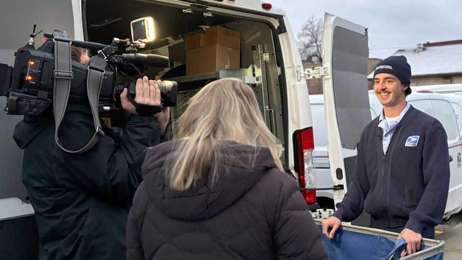 A smiling man wearing a Postal Service uniform stands next to a USPS delivery vehicle and addresses a TV reporter and cameraman