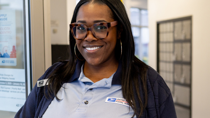 A woman wearing a postal uniform flashes a bright smile while standing in a Post Office retail lobby