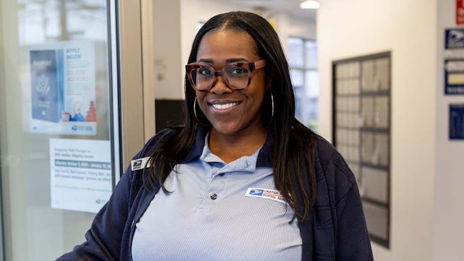 A woman wearing a postal uniform flashes a bright smile while standing in a Post Office retail lobby