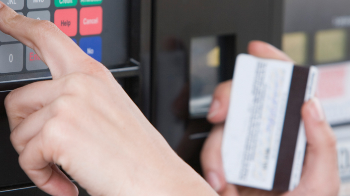 A woman’s hand presses a keypad near a fuel pump; she holds a credit card in her other hand