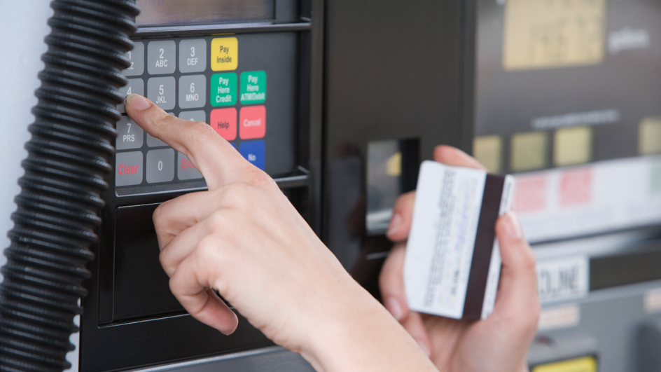 A woman’s hand presses a keypad near a fuel pump; she holds a credit card in her other hand