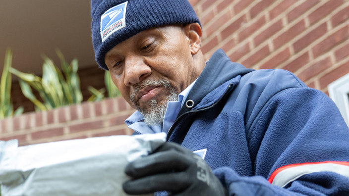 A man wearing a postal uniform examines an envelope near a building decorated for the holidays