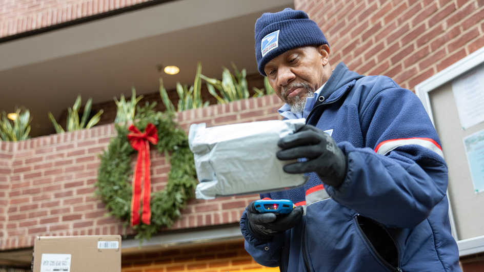 A man wearing a postal uniform examines an envelope near a building decorated for the holidays