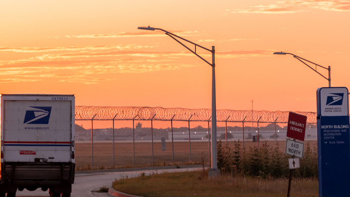A USPS delivery vehicle drives into a sunset