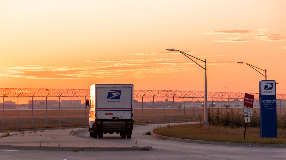 A USPS delivery vehicle drives into a sunset
