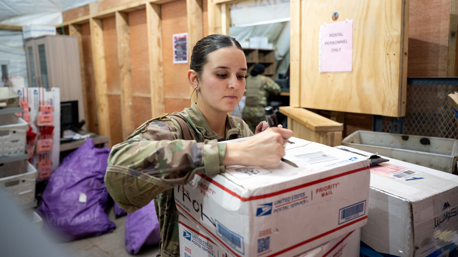 A woman in a military uniform examines a package with USPS branding