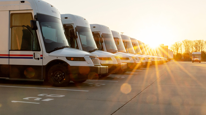 A row of USPS delivery vehicles shown on a parking lot at sunrise