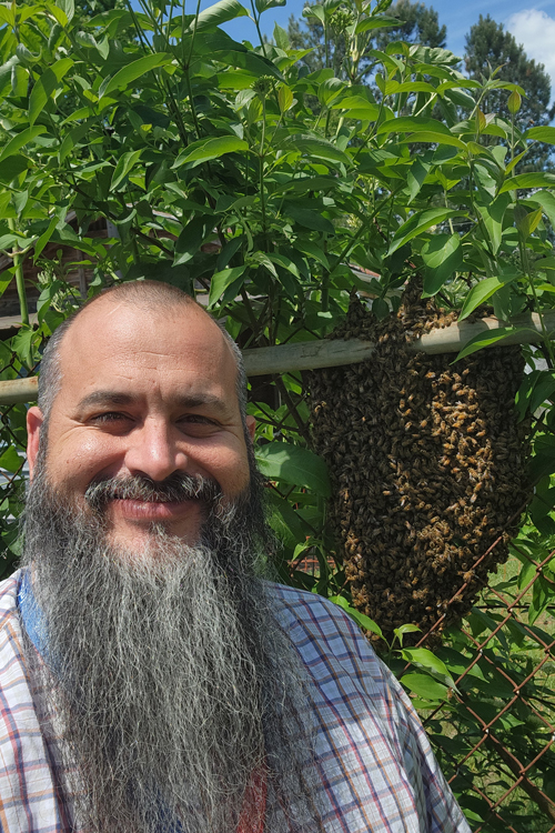 A man smiles whiles standing next to a beehive