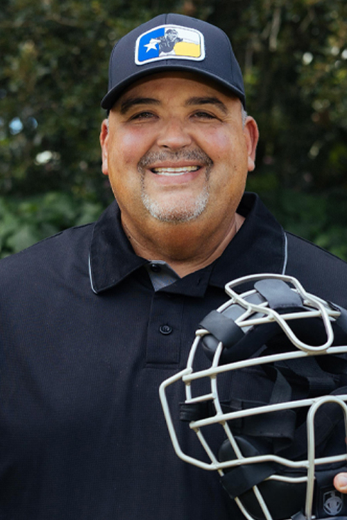 A man wearing a baseball cap and umpire gear stands in a field and smiles