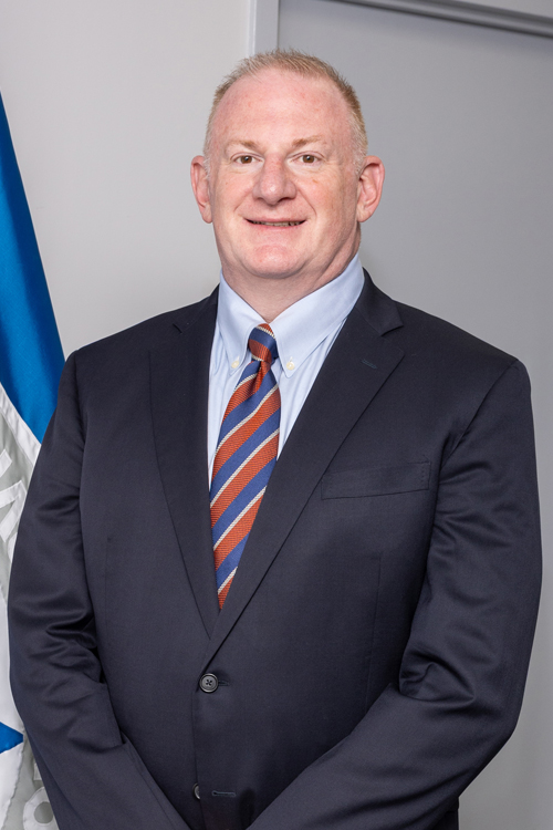 A man wearing a suit stands next to a flag