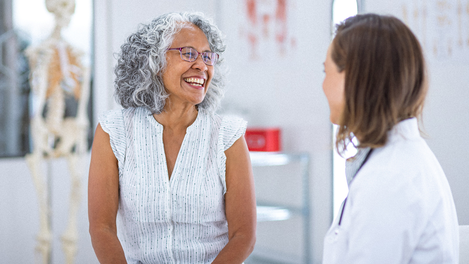A woman sits in an examining room, smiling and speaking with a doctor
