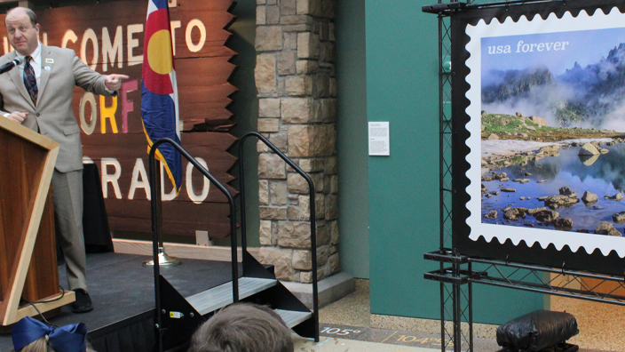 A man behind a podium points to an enlarged image of a postage stamp to his right.