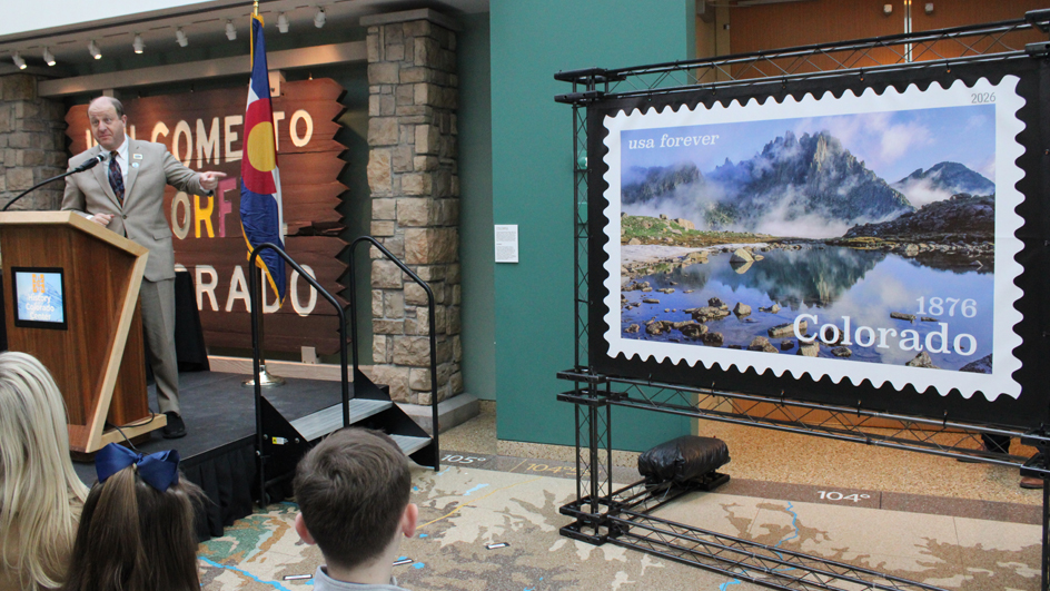 A man behind a podium points to an enlarged image of a postage stamp to his right.