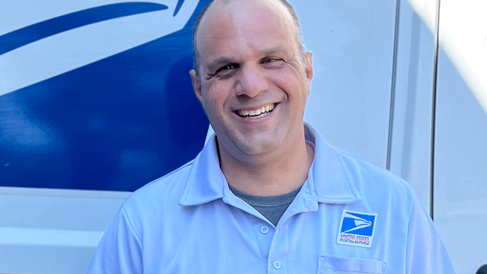 A man in a postal uniform smiles and stands next to a delivery vehicle
