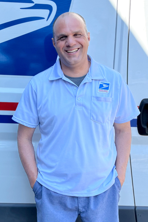 A man in a postal uniform smiles and stands next to a delivery vehicle