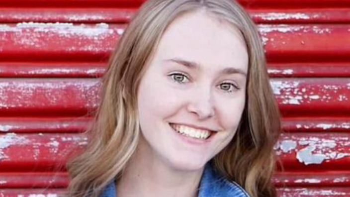 A smiling young woman in a denim jacket stands against a red wall in a rural setting