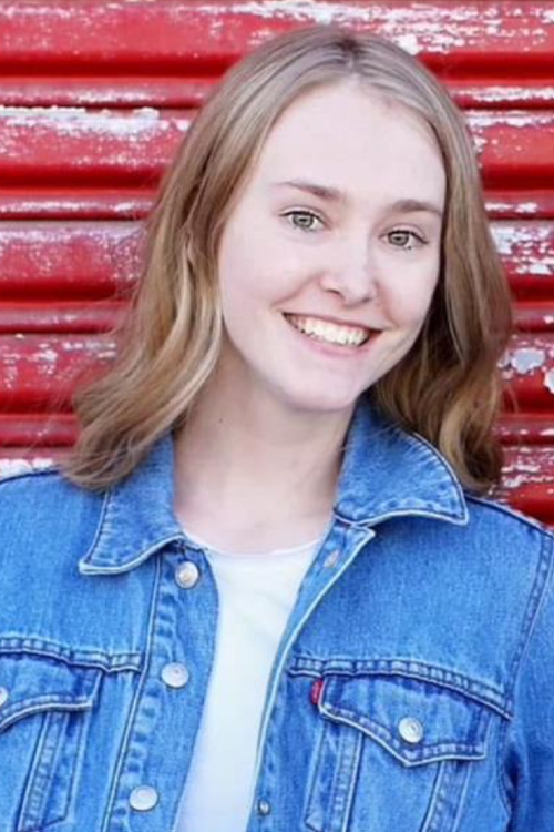 A smiling young woman in a denim jacket stands against a red wall in a rural setting