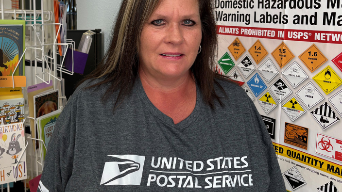 A woman wearing a USPS shirt stands in a Post Office workroom
