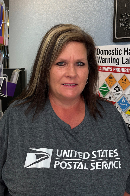 A woman wearing a USPS shirt stands in a Post Office workroom