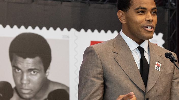 A man in a brown suit and black tie speaks from behind a lectern.