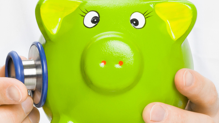 A doctor in a white lab coat uses a stethoscope to listen to a green piggy bank.