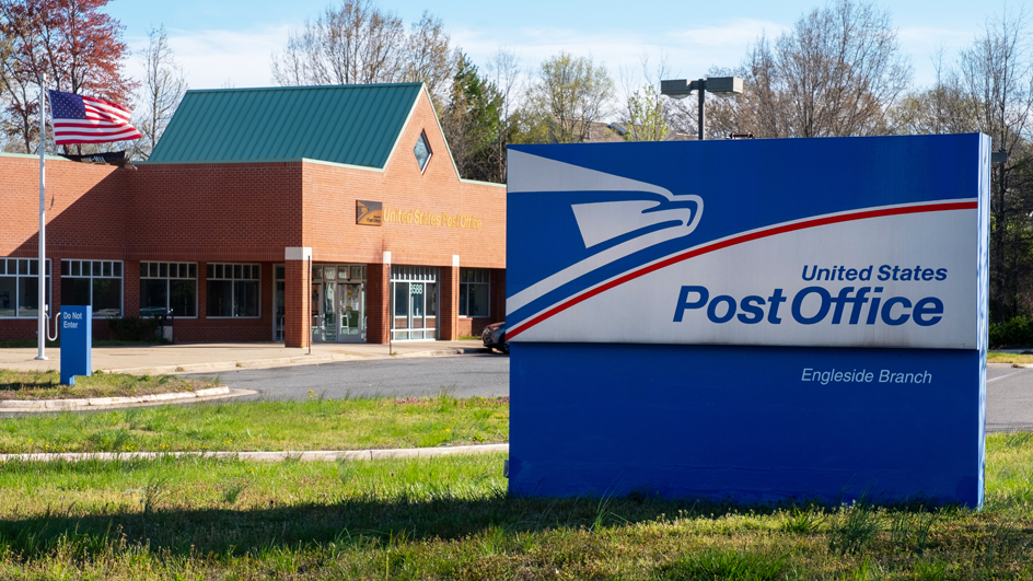 A blue-and-white Post Office sign in the foreground with a brick Post Office building in the background