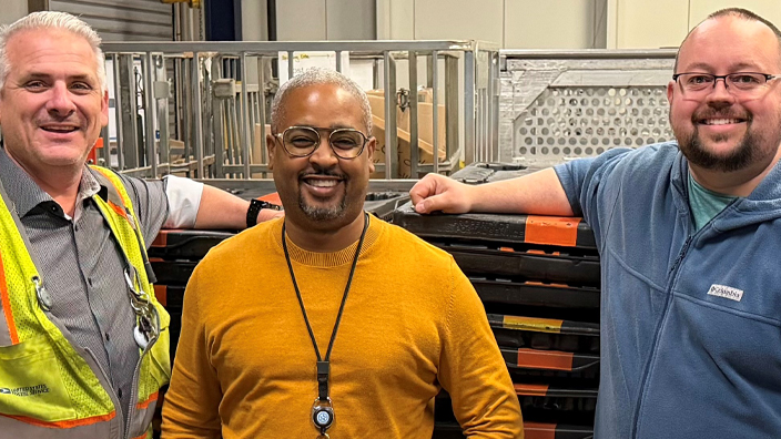 Three men stand in front of a stack of plastic pallets
