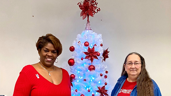 A woman wearing a red dress and a woman wearing blue jeans and a red shirt stand on either side of table with Christmas decorations.