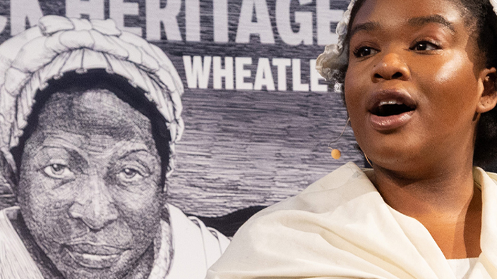 A woman on stage in a Colonial-era costume standing in front of an enlarged stamp image