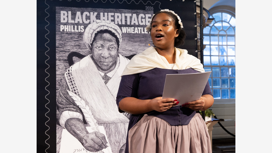 A woman on stage in a Colonial-era costume standing in front of an enlarged stamp image