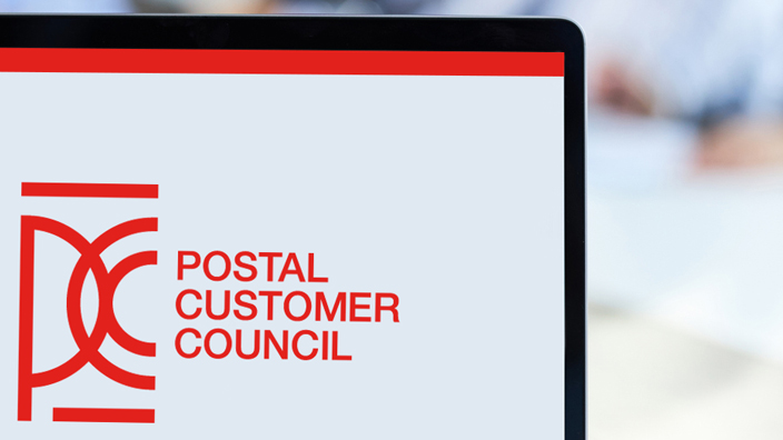 Postal customer council logo on a laptop screen with a blurred background of five people sitting around a table.