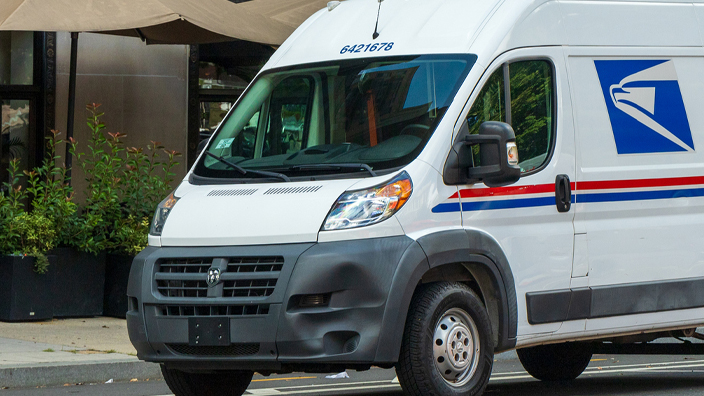 Delivery van on the road in front of a car with a large building and trees in the background.