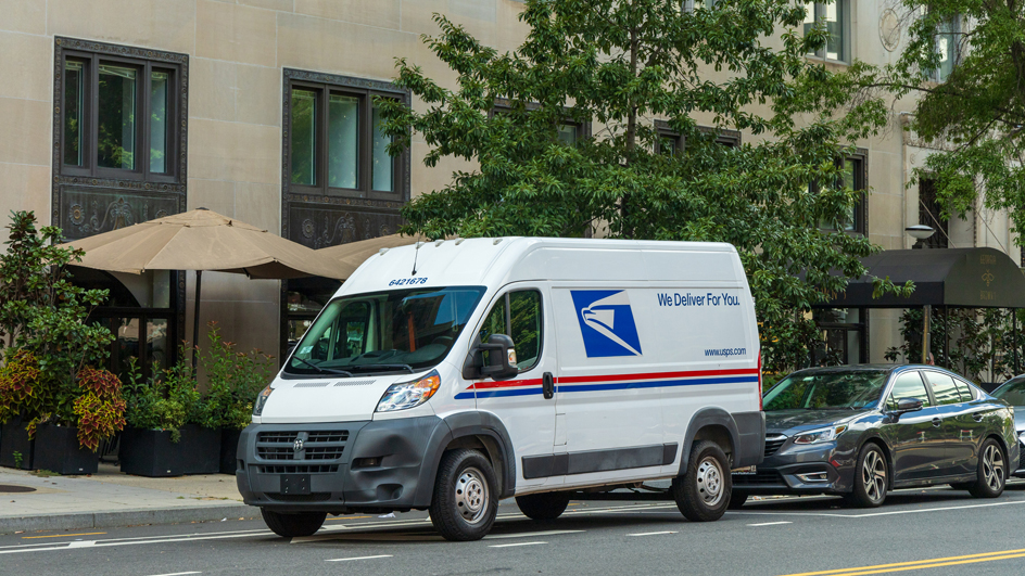 Delivery van on the road in front of a car with a large building and trees in the background.