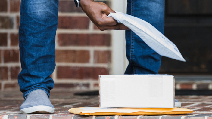 A person wearing blue jeans bending over to pick up packages on the front porch of a house.