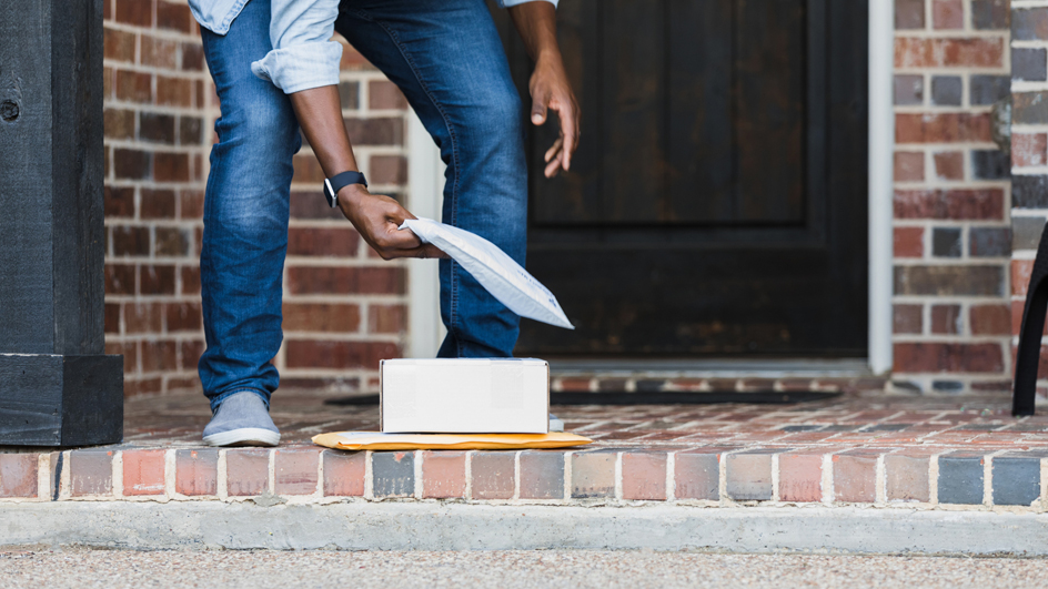 A person wearing blue jeans bending over to pick up packages on the front porch of a house.