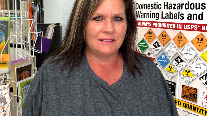A woman wearing a USPS shirt stands in a Post Office workroom