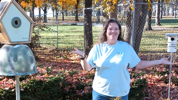 A smiling woman stands in a yard next to a birdhouse