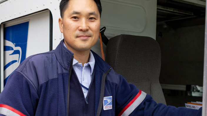 A man wearing a Postal Service uniform stands next to a USPS delivery vehicle