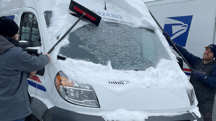 Two men remove snow from the windshield and roof of a delivery van