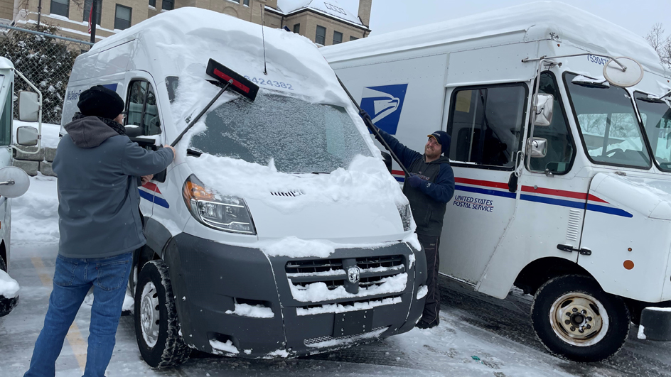Two men remove snow from the windshield and roof of a delivery van