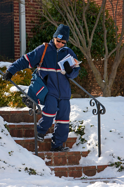 A letter carrier in a blue winter uniform holds onto a handrail while walking down some steps.