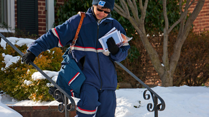 A letter carrier in a blue winter uniform holds onto a handrail while walking down some steps.