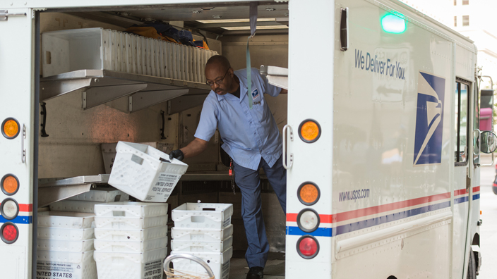 A male USPS employee standing in the back of a delivery truck stacking empty mail bins