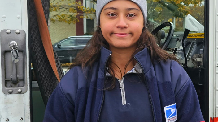 A woman in a postal uniform stands next to a postal delivery vehicle